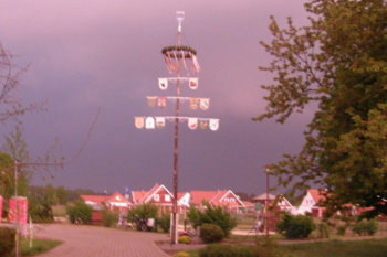Ein Maibaum mit Wappen und Zeichen, der in einer dörflichen Szenerie bei drohendem Gewitterhimmel steht, wobei die aufziehende Dunkelheit der Wolken einen dramatischen Kontrast zum hellen Vordergrund bildet.